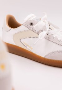 Vit sneaker tillverkad av läder och textil, med en ljus beige accent, platta vita snören och en sula av gummigummi med logodetalj.