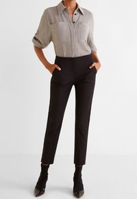 Camicia grigia con bottoni e due tasche sul petto, pantaloni neri su misura e eleganti décolleté neri a punta. Texture morbide e design aderente.