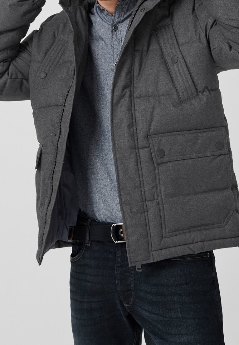 Giacca imbottita grigia con cappuccio, caratterizzata da un design trapuntato, bottoni a scatto e tasche laterali. Indossata sopra una camicia azzurro chiaro a bottoni e jeans.