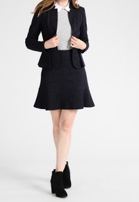 Laivastonsininen pinnaraidallinen takki- ja hameasu, joka sisältää istuvan bleiserin, kellotetun hameen ja harmaan neuleen valkoisella kauluksella. Mustat nilkkurit.