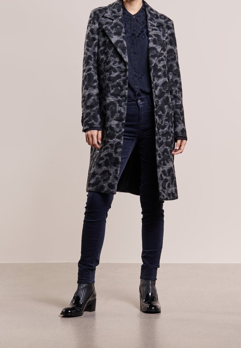 Lang, grijs luipaardprint jas met een notch-kraag, eenrijige knopen, gedragen boven een donker shirt en marineblauwe broek, gecombineerd met glanzende zwarte schoenen.