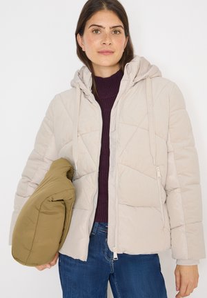 Donna che indossa una giacca a vento beige chiaro sopra un maglione viola e jeans blu, con una giacca piegata verde oliva nella mano sinistra.