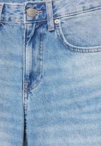 Jean en denim bleu clair montrant la ceinture, le bouton métallique, la boucle de ceinture et la poche avant avec des coutures détaillées.