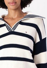 Nainen, jolla on päällä valkoinen neulepaita, jossa on leveät tummansiniset raidat ja leveä kaulus. Paidassa on pieni brodeerattu lippulogotunnus.