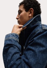 Chaqueta de mezclilla en azul oscuro, con cuello levantado, mangas ajustadas y una textura suave. Costuras visibles y detalles en los puños.