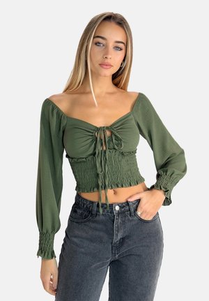 Top corto verde oliva con maniche a sbuffo, vita arricciata e lacci anteriori. Tessuto morbido con bordi arricciati, abbinato a jeans neri a vita alta.