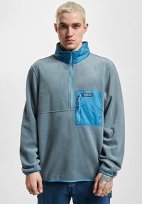 Giovane con capelli corti biondi che indossa un pullover di pile grigio con una tasca a zip blu sul petto e bordi blu, in piedi su uno sfondo bianco.
