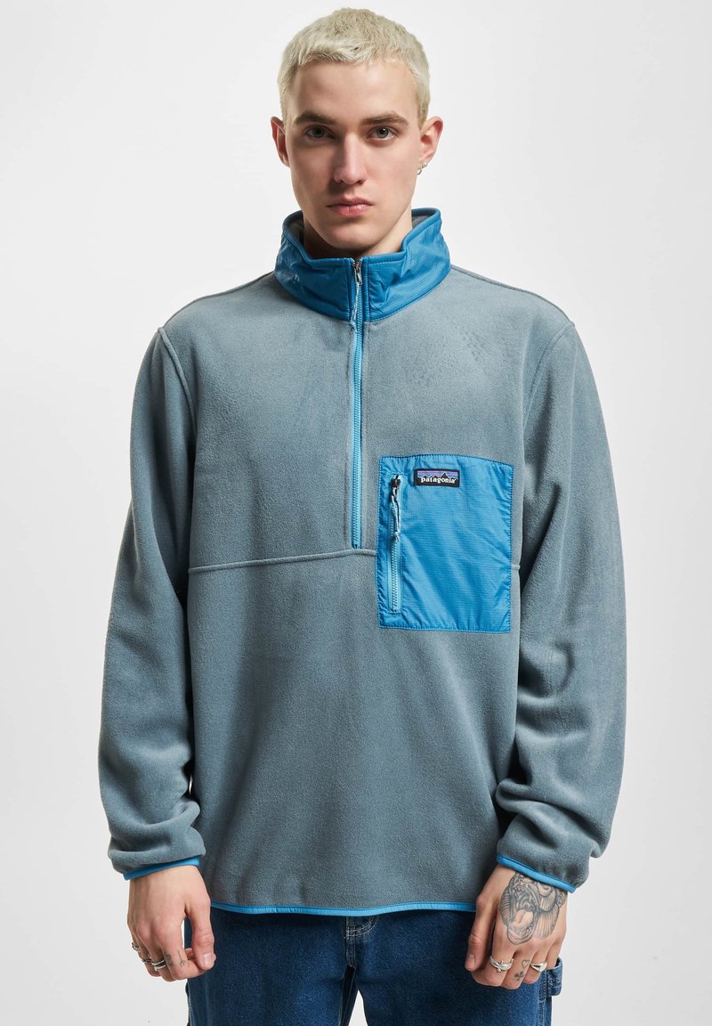 Giovane con capelli corti biondi che indossa un pullover di pile grigio con una tasca a zip blu sul petto e bordi blu, in piedi su uno sfondo bianco.