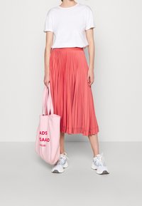 Gonna midi plissettata color corallo, abbinata a una maglietta bianca e sneaker bianche. Una borsa a tracolla rosa con testo rosso è portata dalla modella.