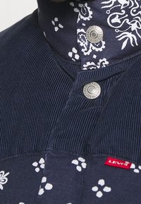 Marinblå skjorta med en texturerad manchesterkrage, prydd med vita blomster- och prickmönster. Har metallknappar och en röd Levi's-tag.