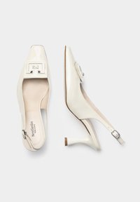 Tacchi slingback in pelle sintetica bianca con punta quadrata, tacco piccolo e una fibbia decorativa sul dorso del piede. Texture liscia con una finitura lucida.