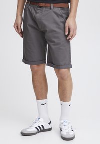 Graue Shorts mit umgeschlagenen Säumen, aus einem glatten Stoff gefertigt. Kombiniert mit einem braunen Gürtel und weißen Socken mit einem schwarzen Logo, getragen mit Sneakers.
