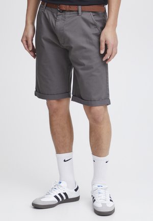 Shorts gris avec des ourlets retroussés, fabriqués en tissu lisse. Associés à une ceinture marron et des chaussettes blanches avec un logo noir, portés avec des baskets.