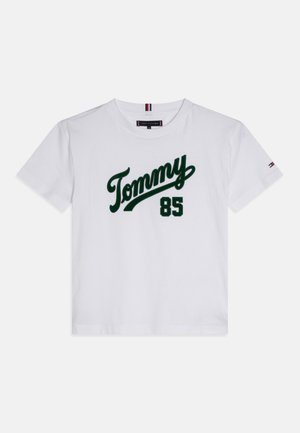 T-shirt blanc à manches courtes avec le script "Tommy" en vert et le numéro "85" à l'avant, comportant un petit logo Tommy Hilfiger sur la manche gauche.