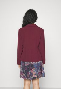 Burgundská přizpůsobená sako s klopovým límcem v kombinaci se vzorovanou modrou sukní, která má květinové a paisley vzory.