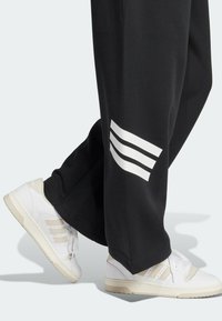 Person som bär svarta byxor med vita ränder och vita sneakers med beige detaljer, står mot en ljus bakgrund.