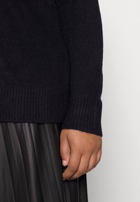 Suéter de punto negro con puños acanalados, combinado con una falda negra con pliegues. Mano visible con textura suave reposando sobre la tela de la falda.