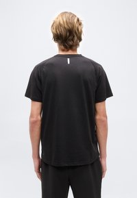 Ξανθός άνθρωπος που στέκεται πλάτη, φορώντας απλό μαύρο κοντομάνικο μπλουζάκι και μαύρο παντελόνι μπροστά σε γκρίζο φόντο.