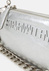 Metalna srebrna torbica s teksturiranom površinom, s "STEVE MADDEN" logotipom u sivoj boji, i sjajnom srebrnom lancem za nošenje.