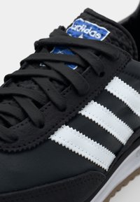 Zwarte athletic schoen met een gladde leren textuur, voorzien van drie witte kartelstrepen, zwarte veters en een blauw Adidas-logo patch.