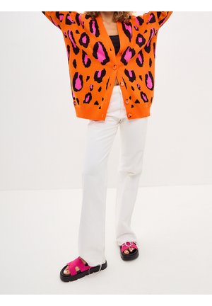 Personne portant un cardigan imprimé léopard orange et rose, un pantalon blanc et des sandales roses, debout devant un fond blanc uni.