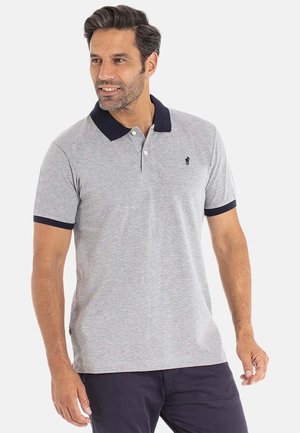 Homme portant un polo gris clair avec un col et des bords de manches foncés, se tenant devant un fond clair uniforme.