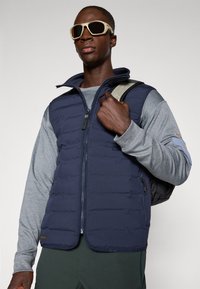 Gilet imbottito blu navy con chiusura a zip, tasche frontali e tessuto testurizzato, indossato sopra una maglietta a maniche lunghe grigia, abbinato a uno zaino nero.