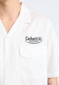 Camisa blanca de manga corta hecha de tela ligera, con cierre de botones, un notable bolsillo frontal y el logo bordado en negro "Carhartt Delicatessen".