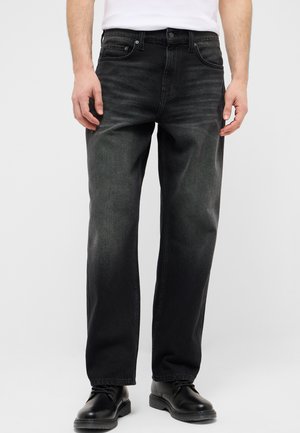 Man wearing black straight-leg jeans with slight fading, paired with black leather shoes and a white shirt, standing against a plain background.