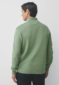 Man met kort donker haar, gekleed in een lichtgroene geribbelde coltrui, gezien van achteren tegen een effen witte achtergrond.