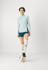 Maglia atletica azzurro chiaro con zip e pannello laterale blu scuro, abbinata a pantaloni corti scuri. Calzini bianchi, scarpe da calcio nere, in piedi su un pallone da calcio multicolore.