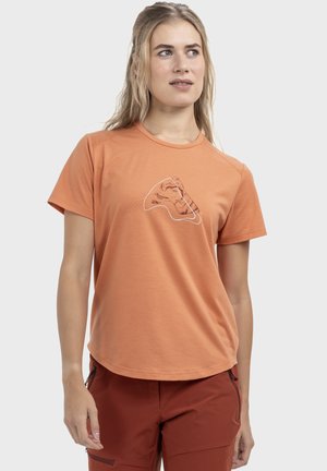 Junge Frau trägt ein orangefarbenes T-Shirt mit Grafik und rostfarbenen Hosen, schaut leicht nach rechts vor schlichtem Hintergrund.