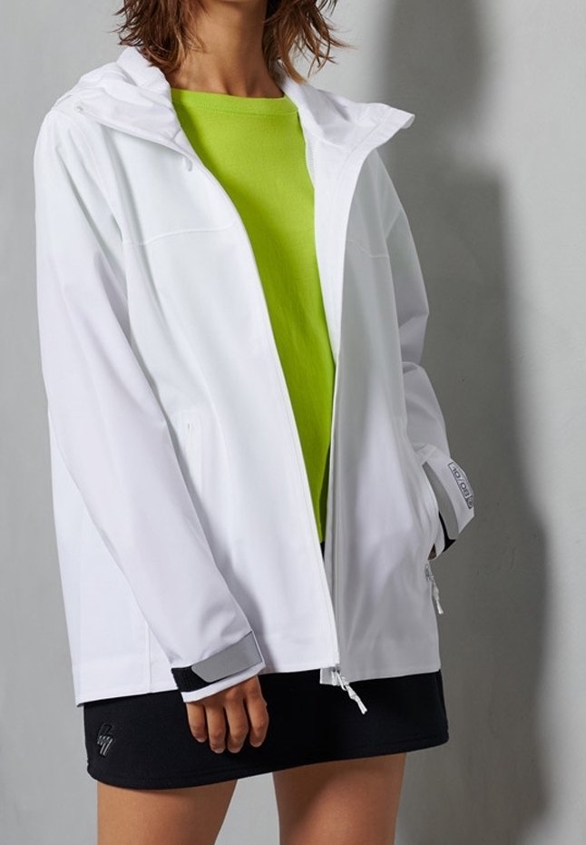 Personne portant une veste à capuche blanche sur un t-shirt vert vif et une jupe noire, debout devant un fond gris uni.