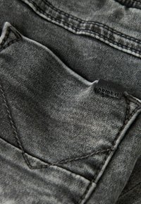 Grå denimstoff med en teksturert finish, med en liten lomme og et svart lærmerke sydd på midjebåndet.