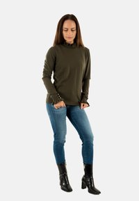Maglietta verde oliva a maniche lunghe con scollatura a volant e dettaglio di bottoni ai polsini, abbinata a jeans skinny blu e stivali neri con tacco.