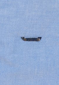 Lichtblauwe textiel met een subtiele textuur, voorzien van een donkerblauw label met een bloemenpatroon dat in het midden is genaaid.