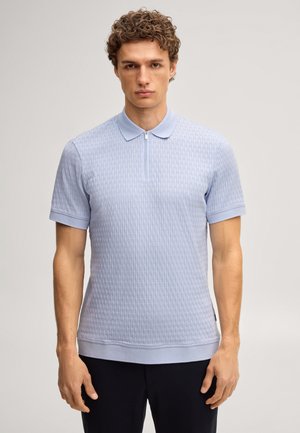 Młody mężczyzna w jasnoniebieskiej teksturowanej koszulce polo z krótkim rękawem i suwakiem, stojący na jednolitym jasnym tle.