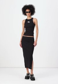Crni rebrasti crop top s logom, uparen s crnom suknjom visokog struka do gležnja. Model nosi crne platforme i sunčane naočale.