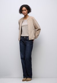 Beige fleece bomberjacka över en ljusbeige topp, kombinerad med mörkblå jeans och bruna ankelstövlar. Enkel, ren design och avslappnad passform.