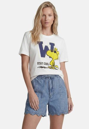Blonde Frau trägt ein weißes T-Shirt mit einem gelben Cartoon-Vogel und dem Text "Stay Cool,", kombiniert mit hellblauen Denim-Shorts mit gewelltem Saum.