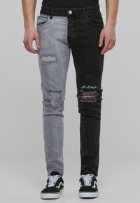 Vaqueros bicolor con una pierna izquierda gris claro y una pierna derecha negra, parches desgatados y el logo bordado "Mr. Knife" en la parte frontal inferior.