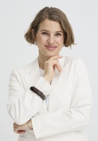 Femme souriante aux cheveux bruns courts portant un blazer blanc, reposant son menton sur la main droite, accessoirisée avec un bracelet foncé et un bracelet chaîne en argent.