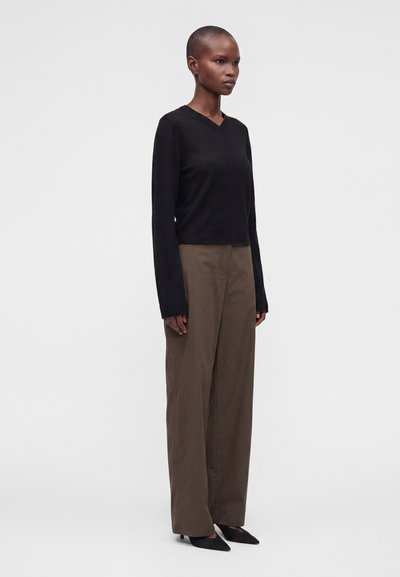 Femme aux cheveux courts portant un haut noir à manches longues, un pantalon large marron et des chaussures noires pointues, debout devant un fond uni.