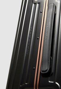 Schwarzer Hartschalen-Koffer mit vertikalen Rillen, kupferfarbenen Akzenten und einem strukturierten schwarzen Ledergriff mit Naht. Markenlogo sichtbar.