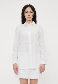 Camicia bianca a maniche lunghe con colletto button-down, realizzata in tessuto leggero; presenta un taglio dritto ed è abbinata a pantaloni corti bianchi.
