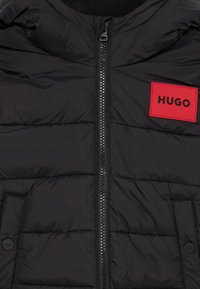 Svart puffer vest med glidelås foran og et rødt rektangulært logopatch merket "HUGO." Har sydd paneler og sidelommer.