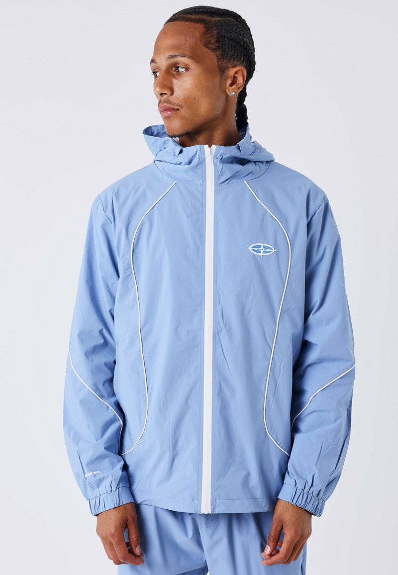Chaqueta cortavientos de color azul claro con cierre de cremallera al frente, detalles de ribete blanco y capucha. Cuenta con puños elásticos y un logo en el lado izquierdo del pecho.