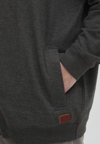 Felpa grigia scuro realizzata in tessuto morbido con tasca a marsupio. Presenta polsini a coste e un'etichetta in pelle marrone all'orlo.