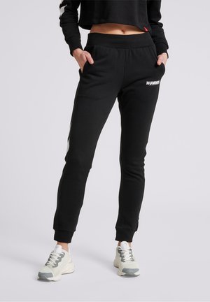 Person iført sorte Hummel joggers med hænderne i lommerne og hvide sneakers, stående mod en lys baggrund.