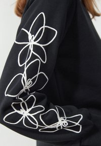 Svart sweatshirt med vita broderade blommönster längs vänster ärm, som visar en jämn textur och detaljerad sömnad.